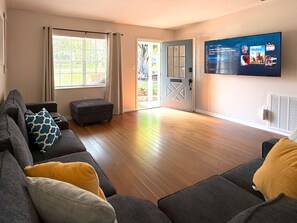 Un salon avec un canapé gris, un pouf et un téléviseur à écran plat fixé au mur.