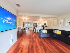 A living room with a flat-screen TV displaying the Disney+ logo, a dining area with a pool table, and a kitchen with white cabinets and a microwave. - Ultimate Family Getaway: 4BR Home with Pool & Games (Winter Haven)