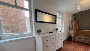 Une pièce avec une commode blanche, un miroir, une plante en pot et une vue sur un bâtiment en briques par la fenêtre.