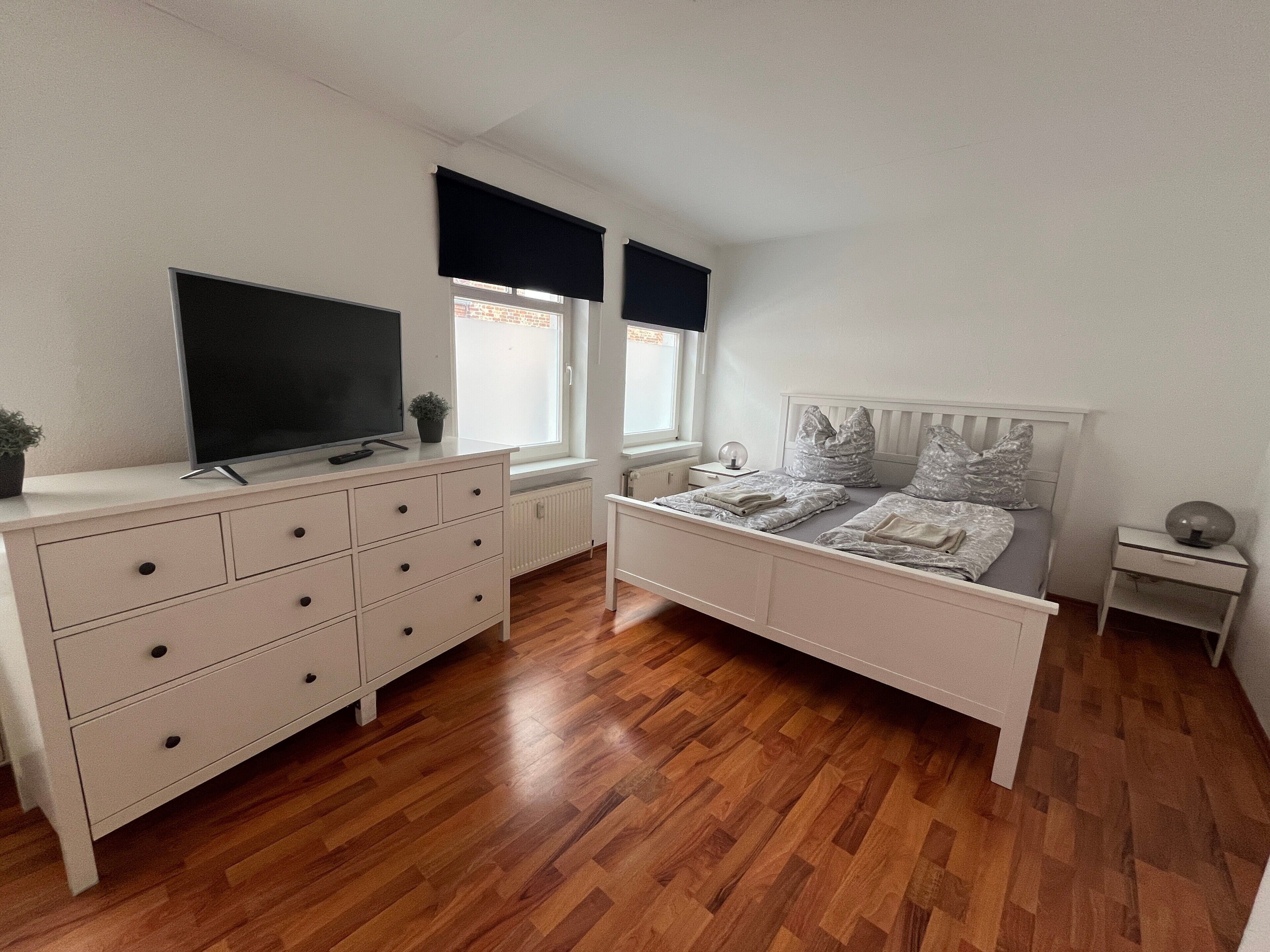 A bedroom with a bed, dresser, TV, windows, and a radiator.
