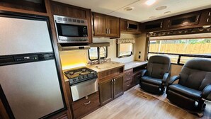 Cocina compacta para autocaravana con estufa, microondas y refrigerador. Hay dos sillas negras y una ventana con vista a una cerca de madera.
