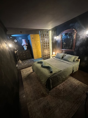 A dimly lit bedroom with a bed, a nightstand, a wall-mounted mirror, and a decorative partition. - Hostal Mustelier (Barcelona)