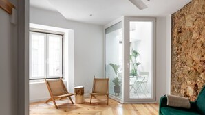 A spacious room with wooden flooring, a green sofa, two wicker chairs, a wooden stool, and a glass door leading to a balcony with plants. - Spacious 6-Bedroom Apt w/ Sunny Terrace in Lisbon (Lisbon)