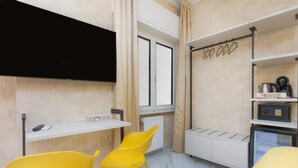 A compact room with a white table, two yellow chairs, a black TV mounted on the wall, a radiator, and a shelf with kitchen items. - Flowery Rainbow (Rome)