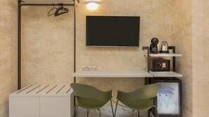 A small, modern room with a white desk, two green chairs, a flat-screen TV, a coffee machine, a mini-fridge, and a wall-mounted heater. - Flowery Rainbow (Rome)