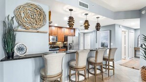A modern kitchen with bar stools, a large decorative wall piece, and pendant lighting. - Cape Harbour Luxury Condo, 2 King Beds, Pool, Shops & Restaurants (Cape Coral)