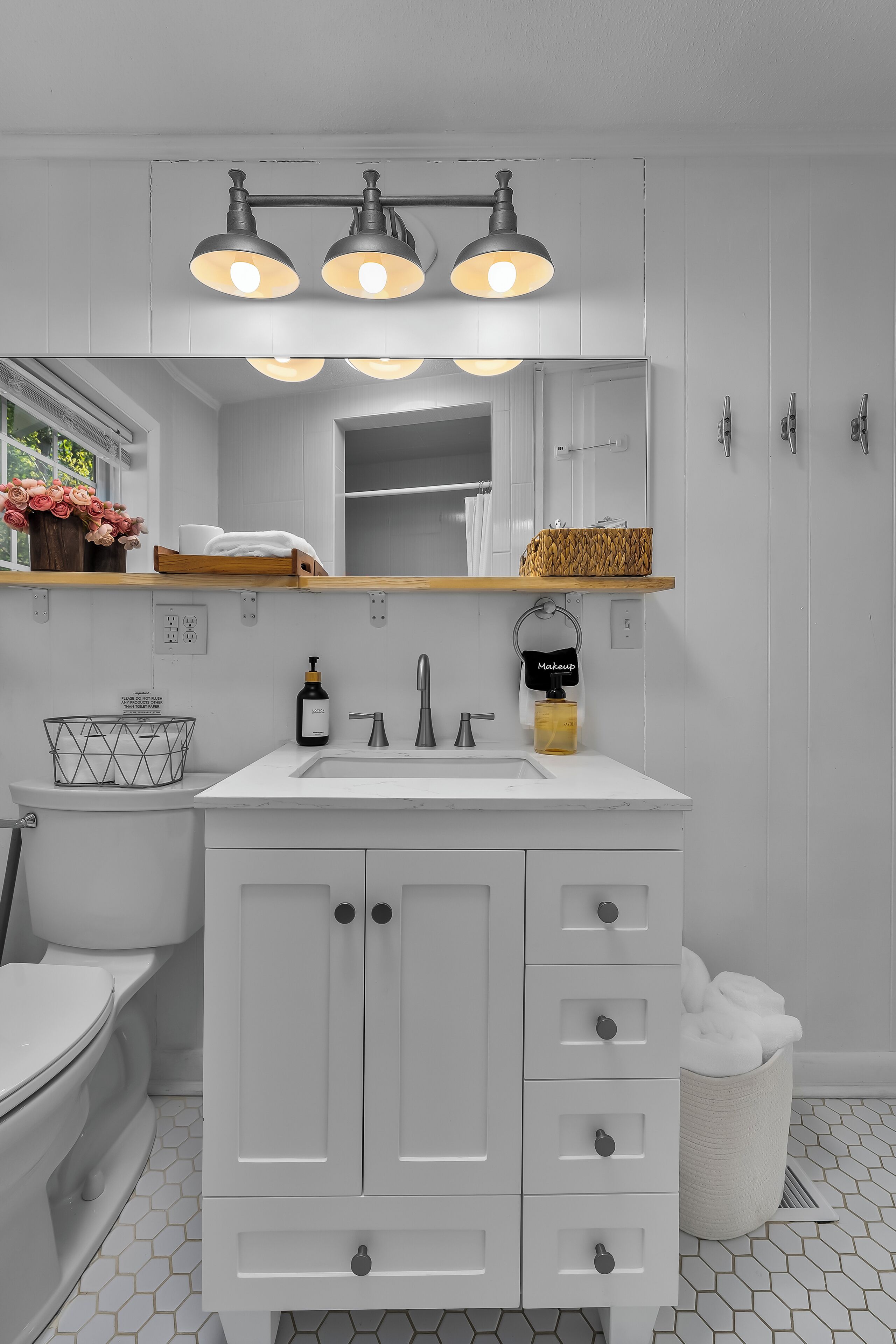 A bathroom with a white vanity, a mirror, a sink, and a shower. There are three lights above the mirror, and a basket of towels is visible.