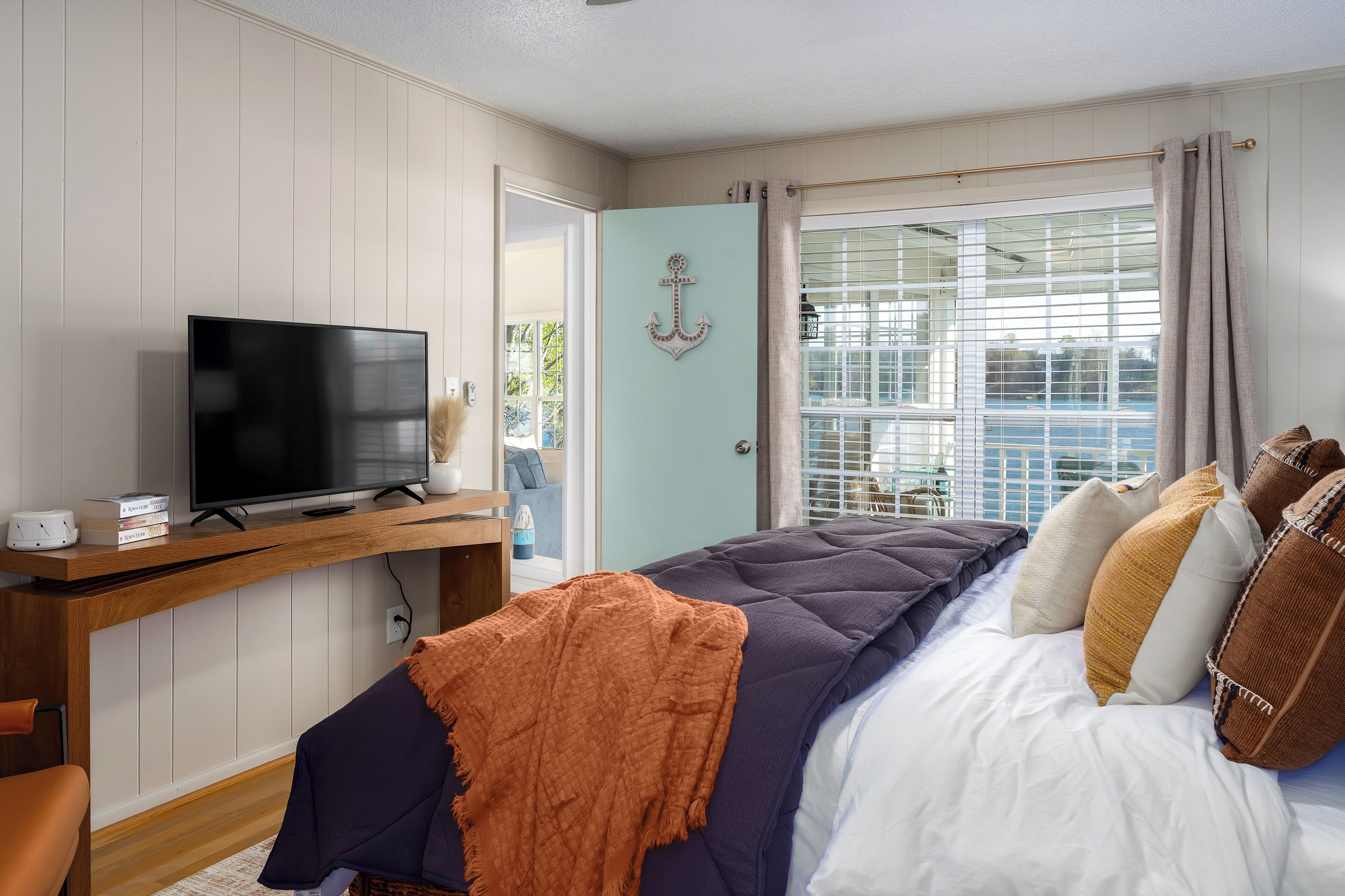 A bedroom with a bed, a television on a wooden stand, a window with curtains, and a door with an anchor decoration.