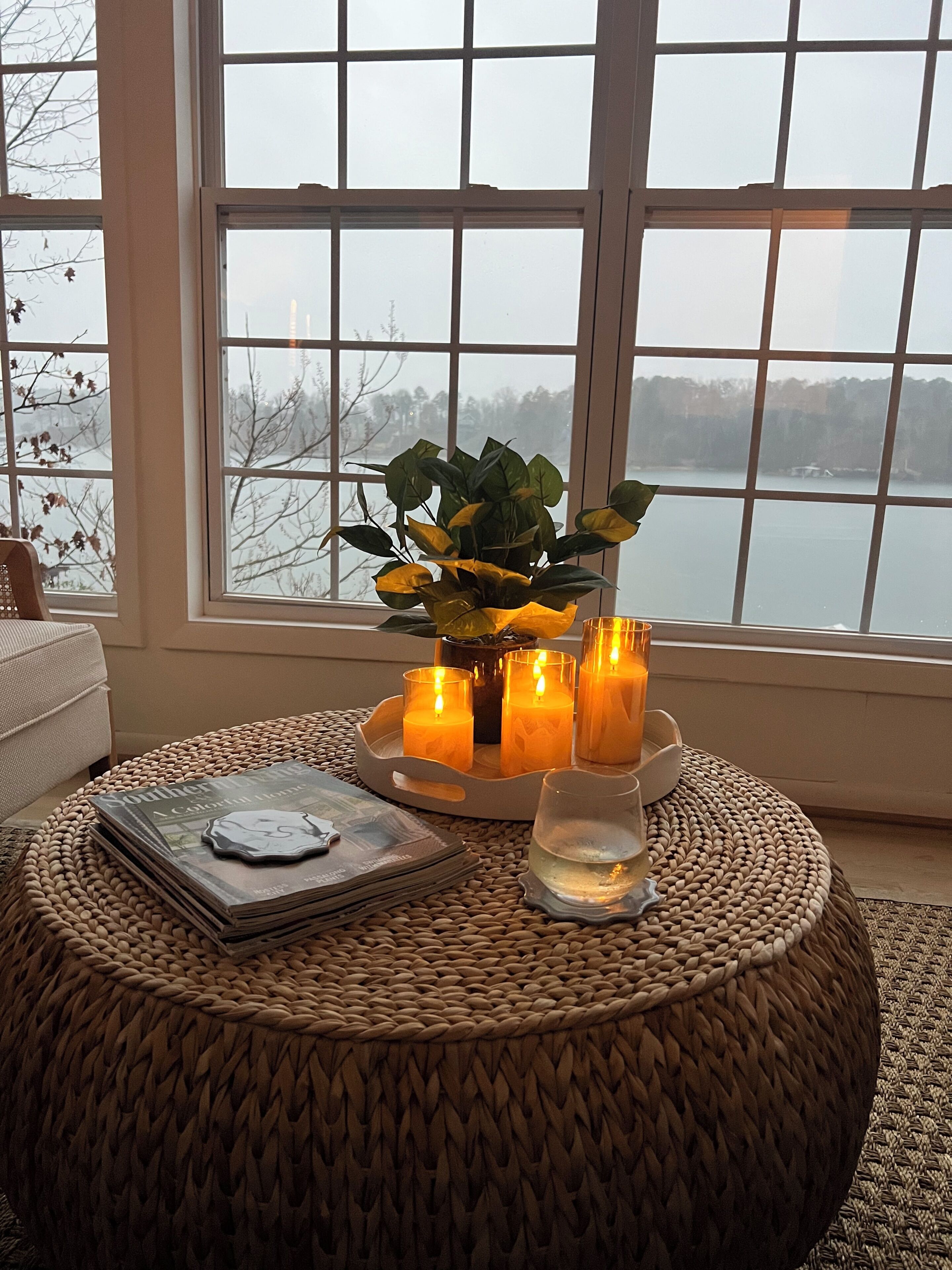 A cozy room with a large window, a woven ottoman, a tray with candles, and a magazine.