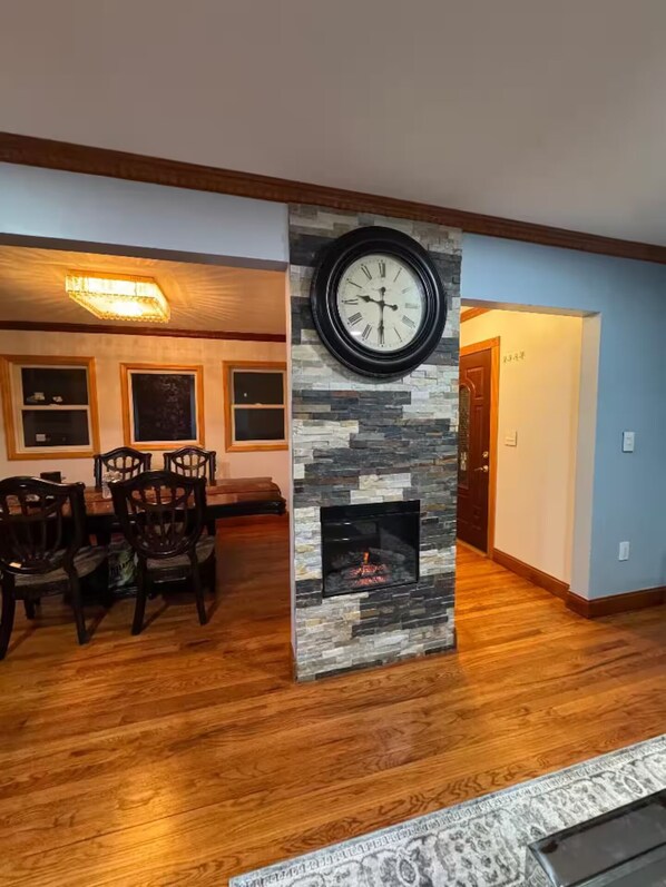 A living room with a stone fireplace, a dining area with wooden chairs, and a large wall clock. - 4-bedroom Entire home in Valley Stream NY, 10 Min to JFK Near Shopping & NYC (Valley Stream)