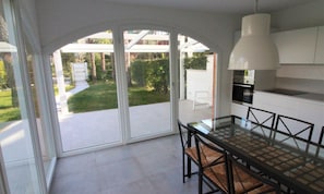A modern kitchen with a glass dining table, white cabinets, and a view of a garden. - Modern Villa a few Steps From the Beach (Lignano Sabbiadoro)