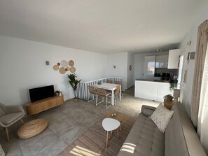 Een moderne woonkamer met een flatscreen televisie, een bank, een eethoek met een witte tafel en een keuken op de achtergrond.