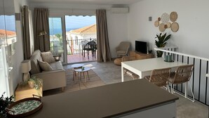 A modern living room with a sofa, a dining table, and chairs, a TV, and a balcony with a view. - Holiday Home 'Los Naranjos' with Shared Pool, Wi-Fi and Air Conditioning (Nerja)