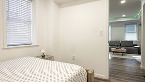 Een slaapkamer met een bed, een klein tafeltje met een vaas en een raam met jaloezieën.