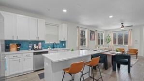 Una cocina moderna con armarios blancos, una isla central con taburetes, una zona de comedor con mesa y sillas, y un ventilador de techo.