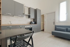 A modern kitchen with grey cabinets, a dining table with chairs, a built-in oven, and a blue sofa in the adjacent living area. - GuestHost - Modern mezzanine studio apartment of 45 m2, ideal for up to 3 people, located on the ground floor of a building (1 step to access).The accommodation is located in the Forlanini area, bordering the large Forlanini Park and the village of Monlu (Milan)