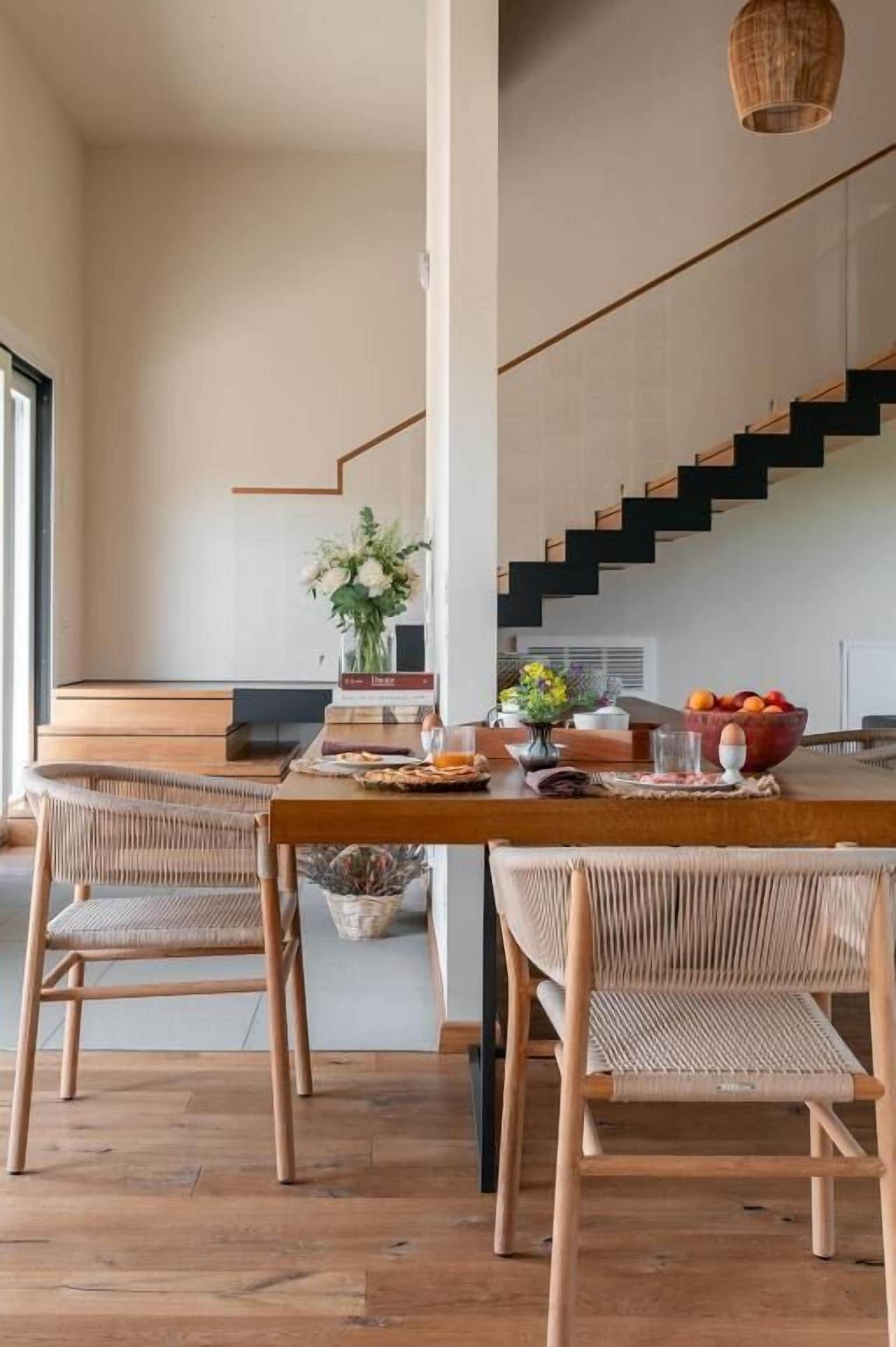 A modern kitchen with a wooden dining table, two wicker chairs, a vase of flowers, and a fruit bowl.