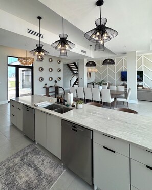 A modern kitchen with a large island, pendant lighting, and a dining area with chairs. - Seaside Villa - Brand New Modern Luxury 7 Bedrooms + 9 Bathrooms + Concierge (Exuma)