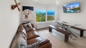 Ein Wohnzimmer mit einer Ledersofa, einem Glastisch, einer Bank, einem Ski-Dekokissen, einem Bergbild und Blick auf einen See. An der Wand ist ein Fernseher angebracht.