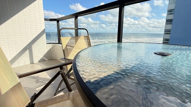 Une table à plateau en verre, une chaise, une vue sur l’océan et un bâtiment à la façade recouverte de carreaux.