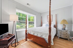 A four-poster bed with a white bedspread, a television on a wooden stand, a lamp on a side table, and a window with blinds. - Lake Norman cottage with stunning views (Mooresville)