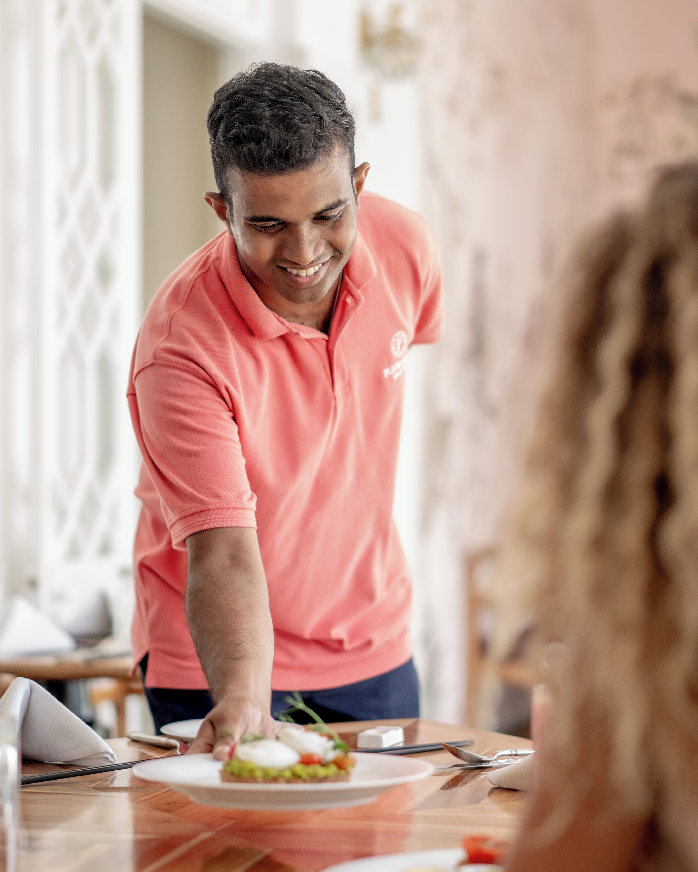 A person in a pink shirt serving food at a table with a white plate, a knife, and a spoon.