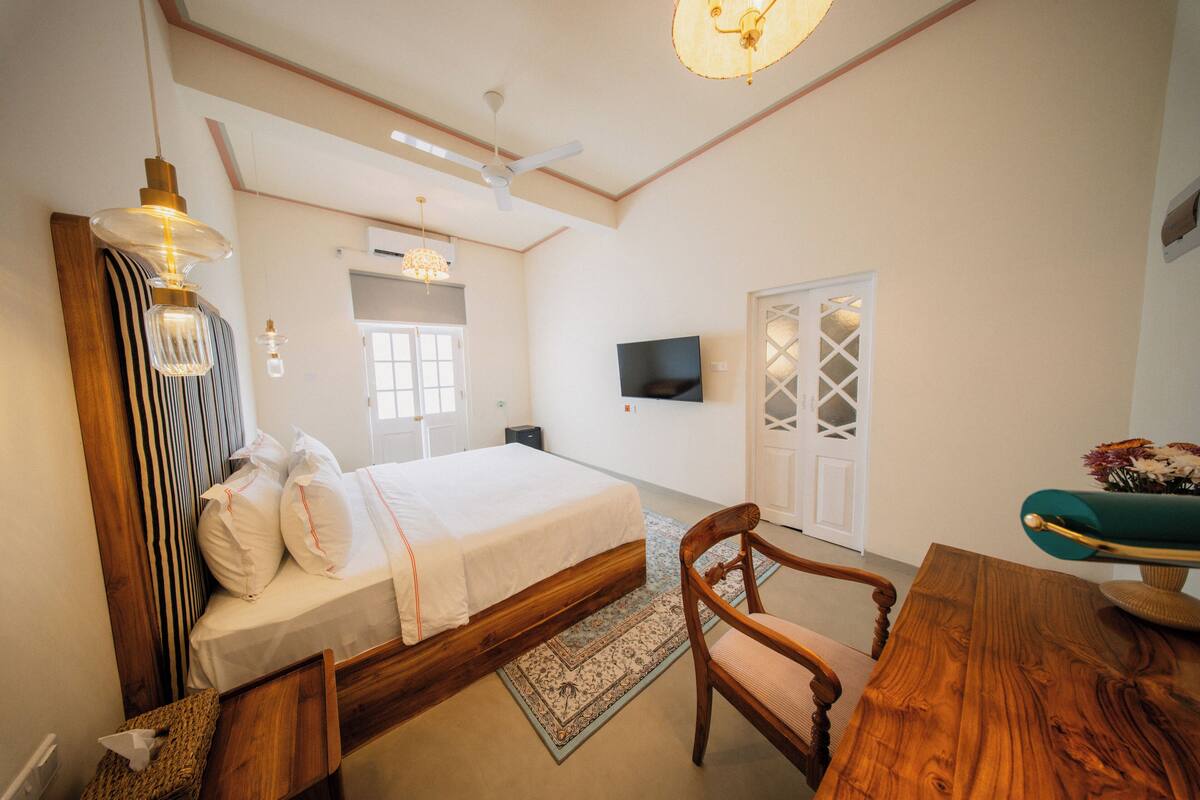 A bedroom with a bed, a wooden chair, a table, a television, and a ceiling fan.