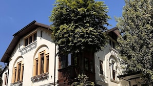 Un edificio de dos pisos con un balcón, un árbol grande en frente y un coche estacionado.