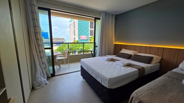 A hotel room with a bed, a small table, a chair, and a balcony with a view of buildings. - Porto Baobas Vista Mar 102 By LP (Ipojuca)
