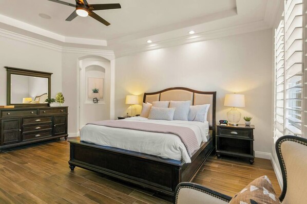 A bedroom with a large bed, dark wooden dresser, two bedside tables with lamps, a ceiling fan, and a large window with blinds. - Boss Mode/Pickleball/Gameroom (Indio)