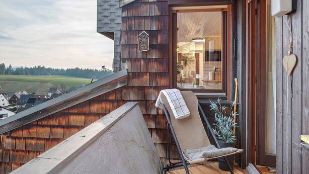 Un balcone in legno con una sedia e una pianta, vista sulla campagna e ingresso di un edificio.