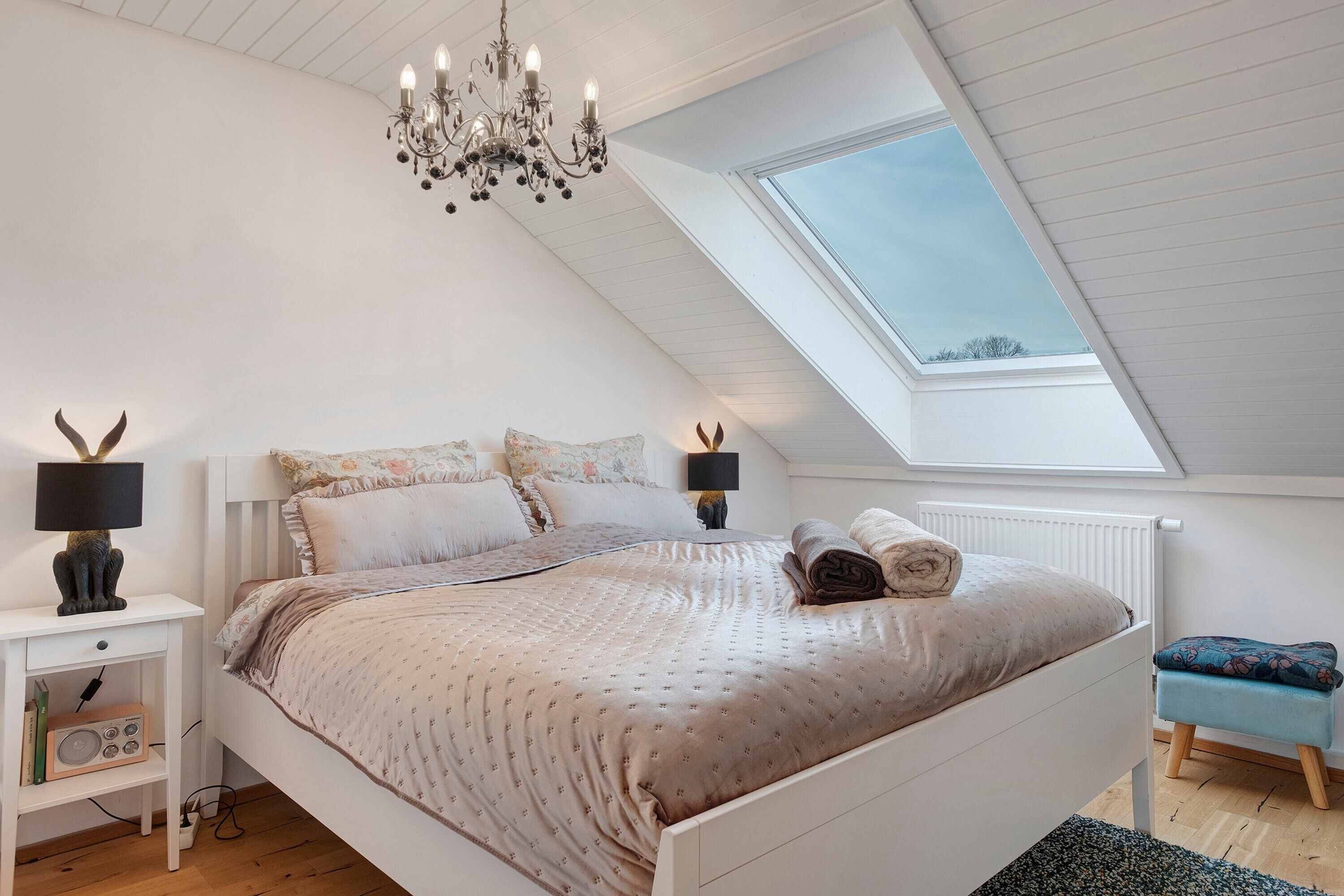 A bedroom with a sloped ceiling, a bed with a patterned bedspread, two bedside lamps, a small table with a radio, and a skylight.