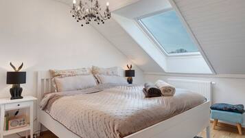 A bedroom with a sloped ceiling, a bed with a patterned bedspread, two bedside lamps, a small table with a radio, and a skylight.