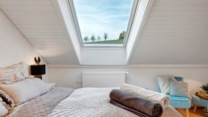 Una camera da letto con una finestra a lucernario, un letto con cuscini, una lampada da comodino, un termosifone e una poltrona blu.