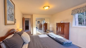 Ein Schlafzimmer mit Bett, einem Holzschrank, einem Fenster mit Vorhängen und einem zur anderen Kammer führenden Flur.