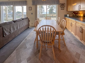 A dining area with a wooden table and chairs, a sofa, and a view through large windows. - Ocean View Retreat on 2 acres (Coos Bay)