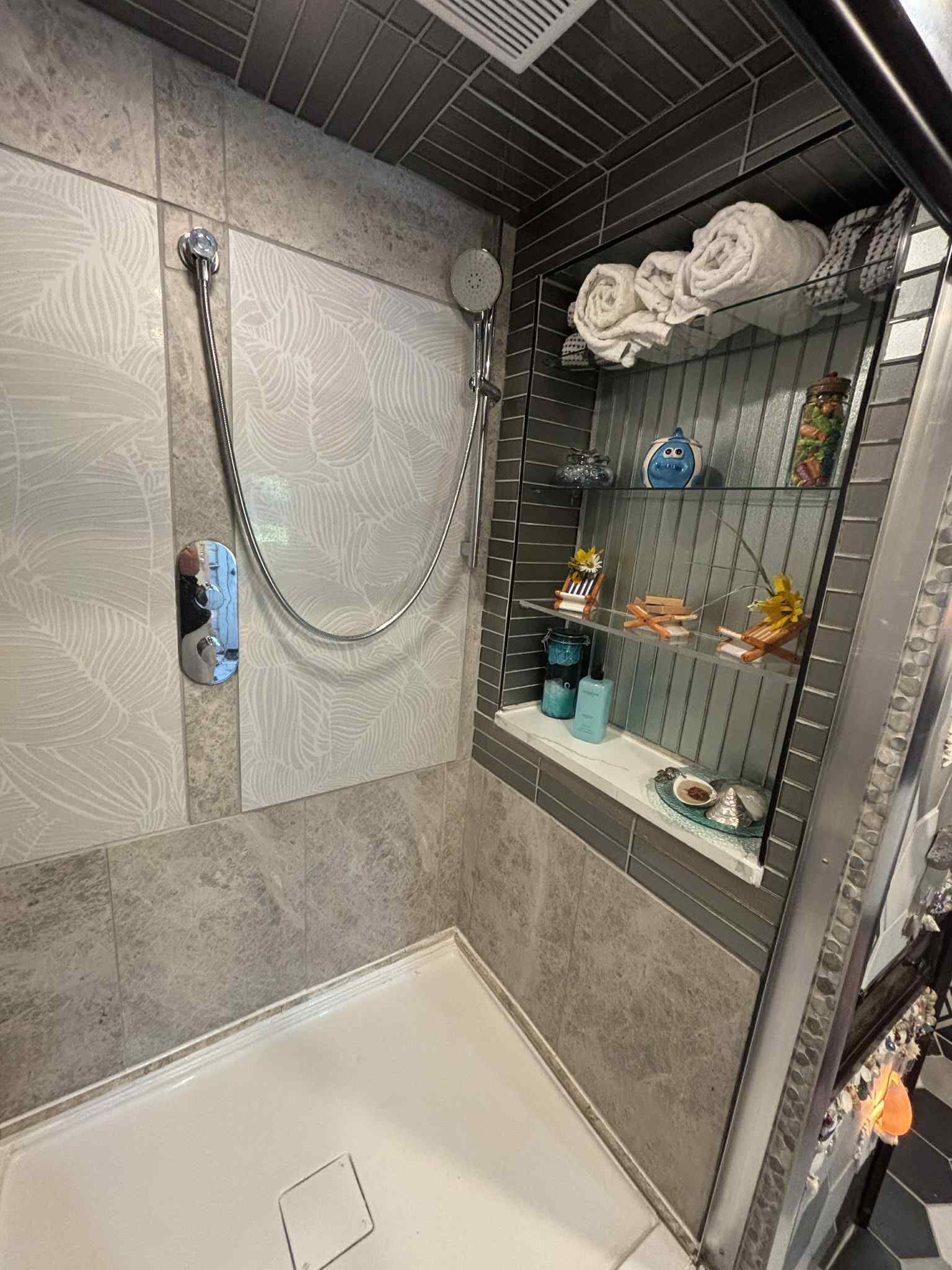 Eine Dusche mit Handbrause, ein Glastrennregal mit Pflegeprodukten und ein Handtuchhalter mit gefalteten Handtüchern.