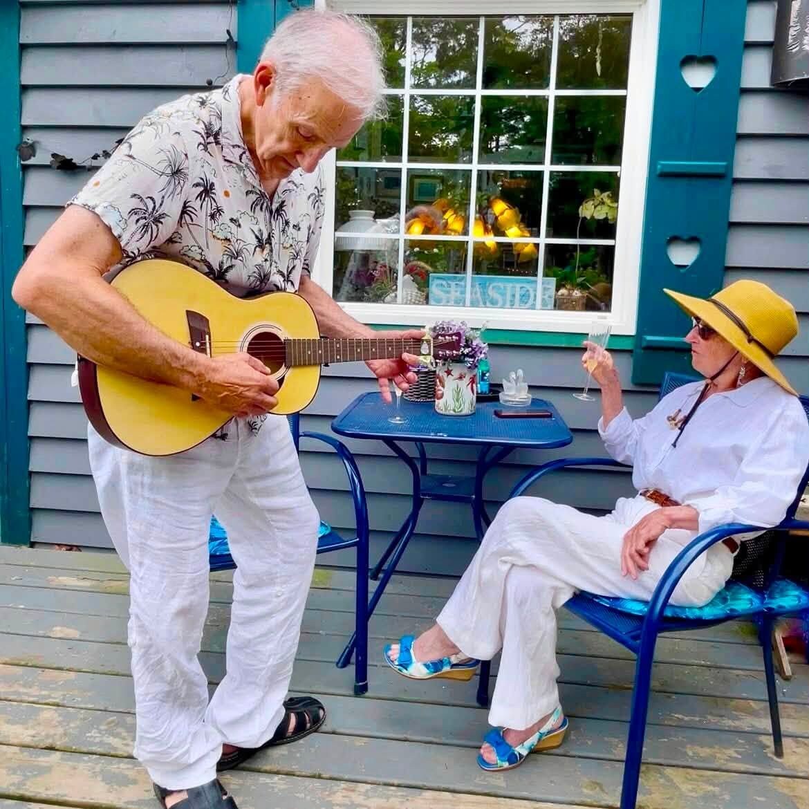 Un homme joue de la guitare sur une véranda où se trouvent une table et des chaises, et une femme est assise à proximité, un verre à la main.