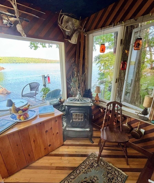 A cozy cabin with a rustic interior, a wooden floor, a stove, a chair, and a view of a lake.