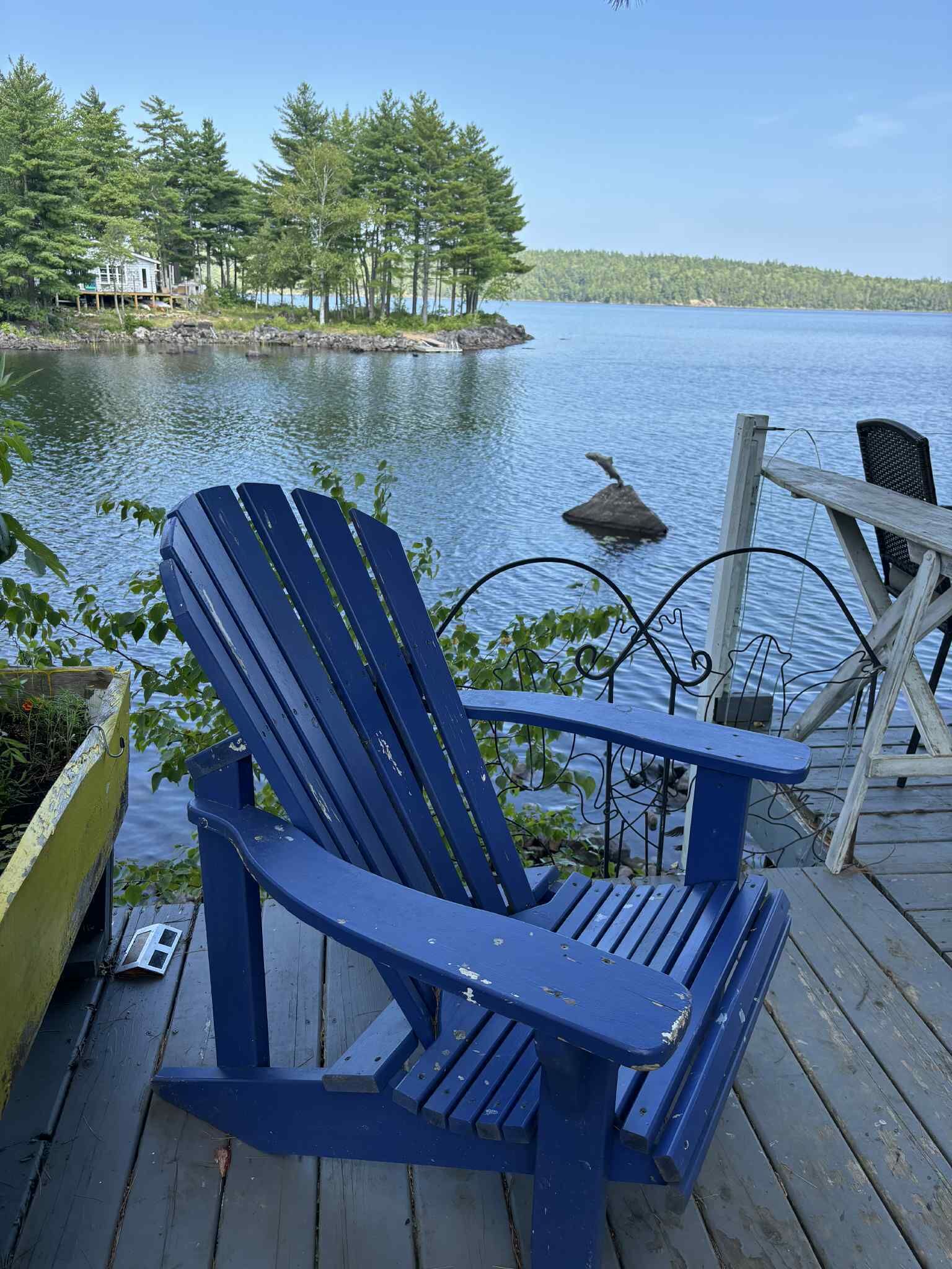 A blue Adirondack chair on a wooden dock overlooking a lake with a small island and trees in the background.