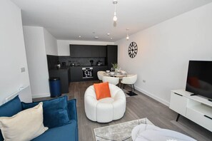 Ein modernes Wohnzimmer mit einer Couch, einem weißen Sessel, einem Essbereich und einer Küche im Hintergrund.