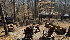 Un camping arbolado con un pozo de fuego, bancos de madera y una cabaña rústica al fondo.