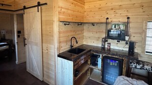 Interior de una cabaña de madera con una zona de cocina, que incluye un lavamanos, refrigerador y una puerta corrediza.