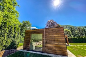 A modern wooden cabin with a glass door, surrounded by lush greenery and a clear blue sky.