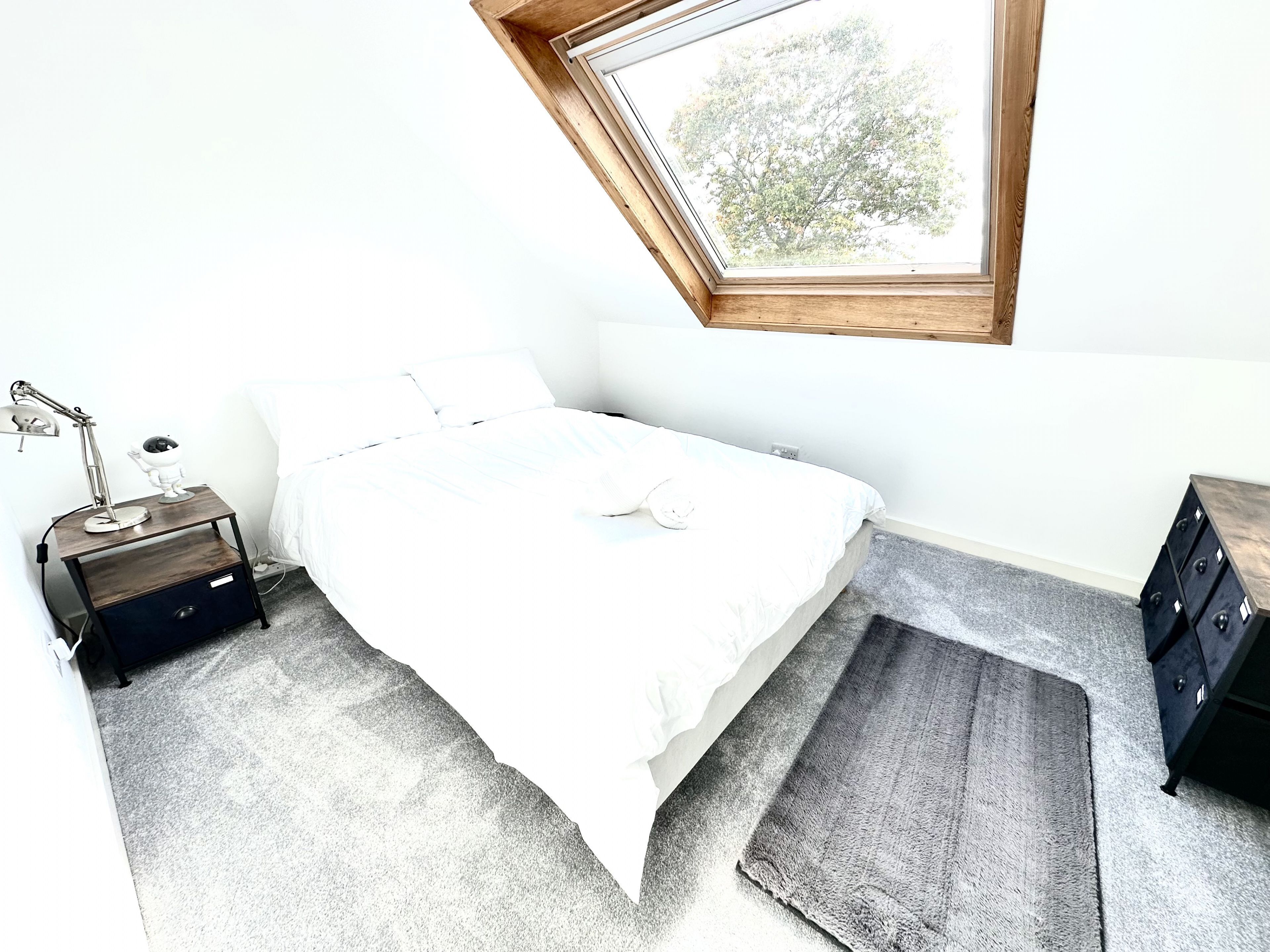 Une chambre à coucher avec un puits de lumière, un lit, une table de chevet avec une lampe, une commode et un tapis.