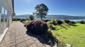 A paved pathway leading to a building with a glass door, a large bush, and a view of a lake with hills in the background. - Spacious Sleeps 10 4 Bedroom Cottage in Inveraray, Fantastic Loch Views and Big Garden (Inveraray)