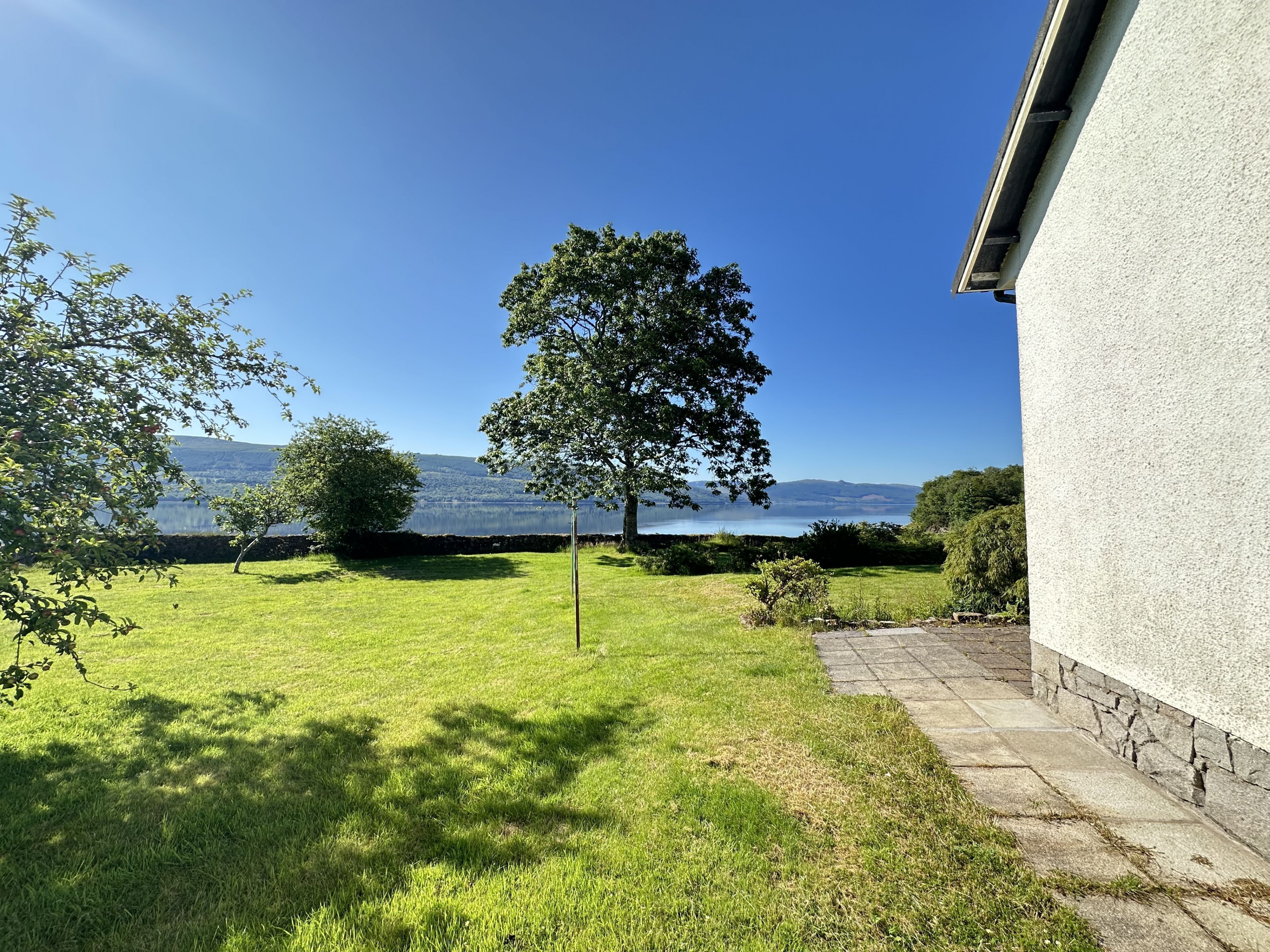 A grassy yard with a tree, a stone wall, and a view of a lake and mountains.
