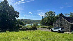A stone building with a car parked in front, a grassy field, and a lake in the background. - Romantic Secluded Cosy Stone Cottage Beside the West Loch, Beautiful Argyll (Tarbert)