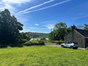 A stone building with a car parked in front, a grassy field, and a lake in the background. - Romantic Secluded Cosy Stone Cottage Beside the West Loch, Beautiful Argyll (Tarbert)
