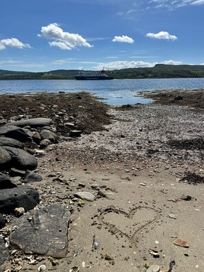 A rocky shoreline with a ship in the distance, a clear sky, and a hilly landscape. - Romantic Secluded Cosy Stone Cottage Beside the West Loch, Beautiful Argyll (Tarbert)